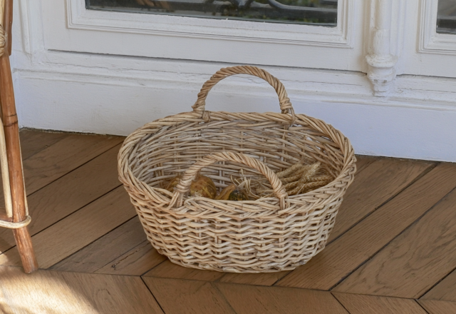 A rustic woven storage basket with double handles sitting on hardwood floors filled with dried wheat.