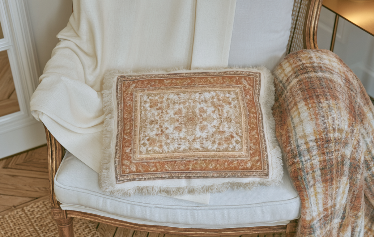 A vintage wooden armchair styled with an ornate fringe accent pillow, a cream throw blanket, and a plaid throw.