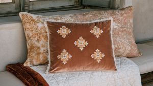 A collection of decorative throw pillows featuring velvet, floral, and paisley prints styled on a quilted neutral bench.
