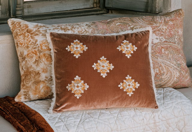 A collection of decorative throw pillows featuring velvet, floral, and paisley prints styled on a quilted neutral bench.