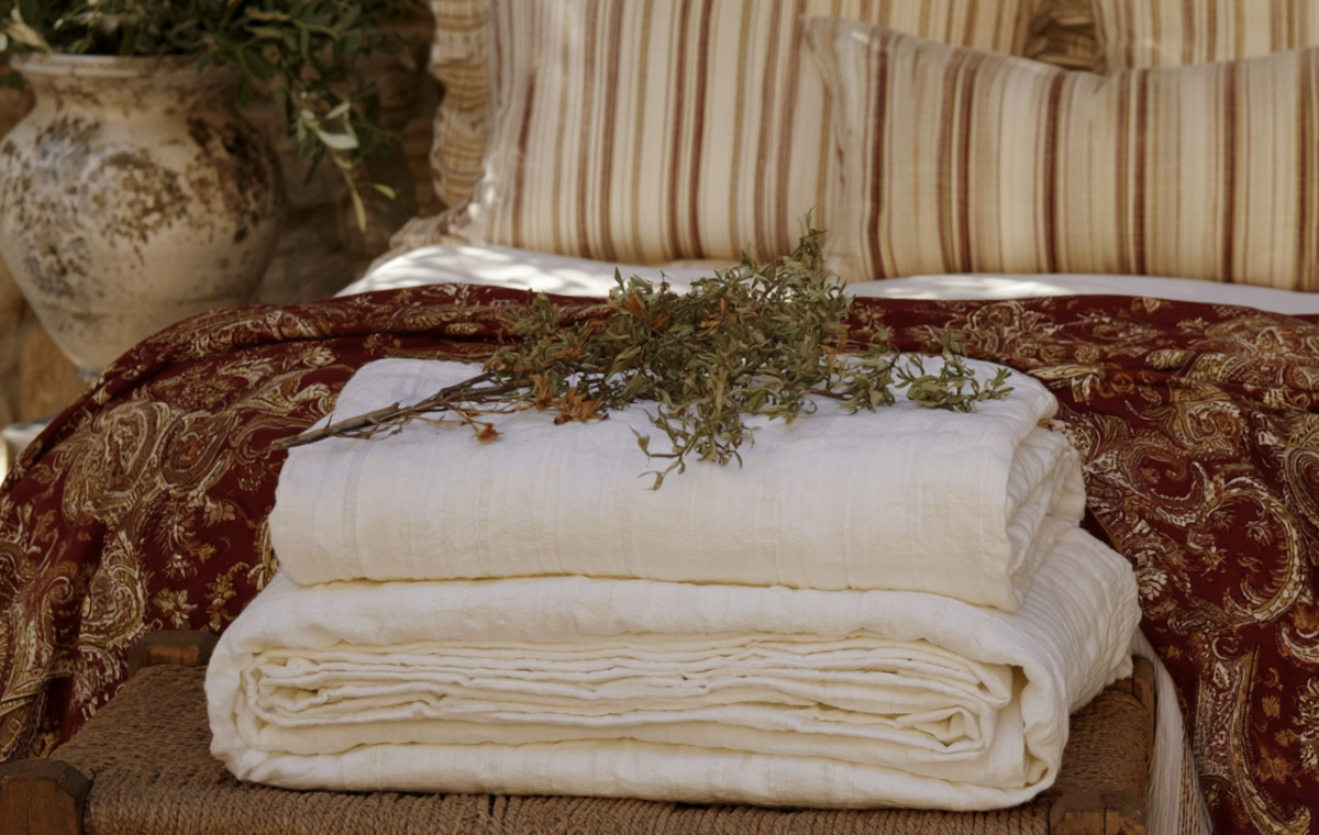Stacked white linen quilts and patterned throw blankets styled on a rustic wooden bench.