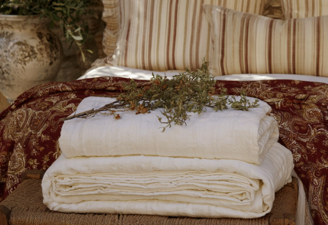 Stacked white linen quilts and patterned throw blankets styled on a rustic wooden bench.