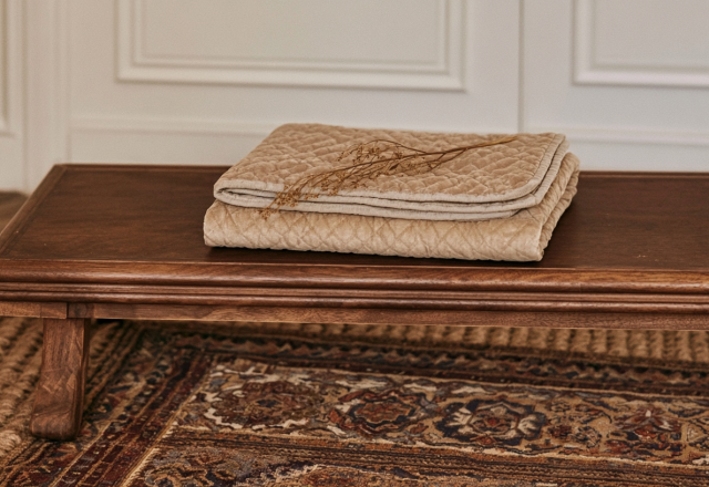 Low wooden coffee table styled with a quilted neutral blanket and dried botanical sprig over a vintage rug.