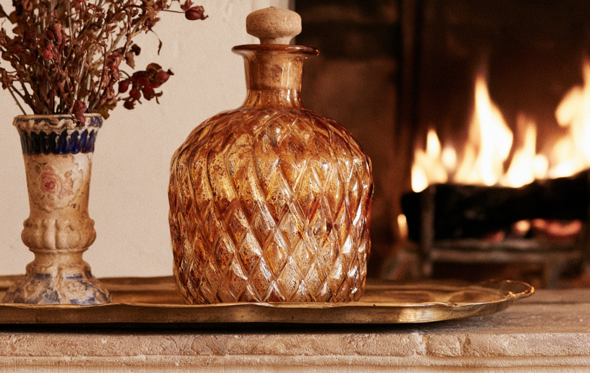 Amber glass decanter and vintage vase styled on a brass tray atop a stone mantelpiece with a fireplace