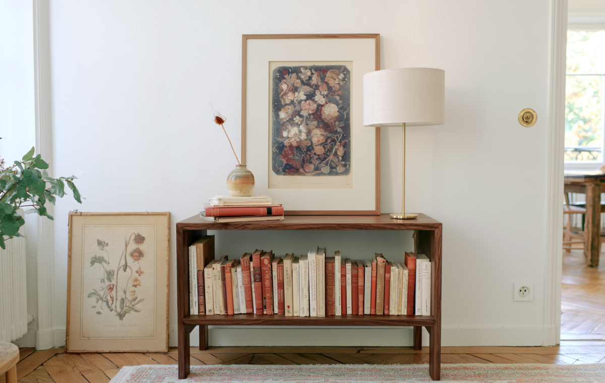 Low wooden bookshelf styled with rows of books, a framed floral print, and a modern brass table lamp in a bright room.