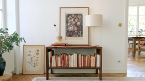 Low wooden bookshelf styled with rows of books, a framed floral print, and a modern brass table lamp in a bright room.