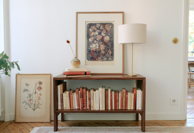 Low wooden bookshelf styled with rows of books, a framed floral print, and a modern brass table lamp in a bright room.
