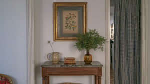Console table styling with marble top, vintage botanical art, and an amber glass vase filled with green stems in a hallway.