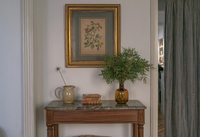 Console table styling with marble top, vintage botanical art, and an amber glass vase filled with green stems in a hallway.