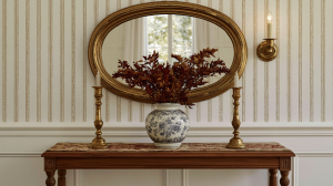 Console table styling featuring an oval gold mirror, vintage brass candleholders, and a blue and white vase in a hallway.