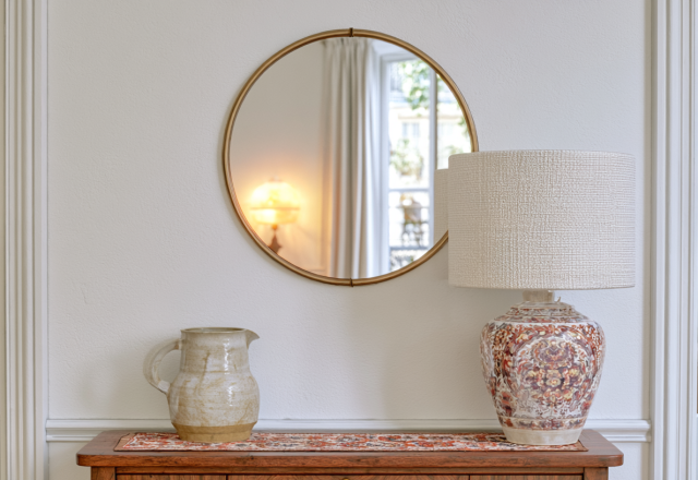 Elegant entryway table styling featuring a round brass mirror, ceramic lamp, and patterned runner on a wooden console.