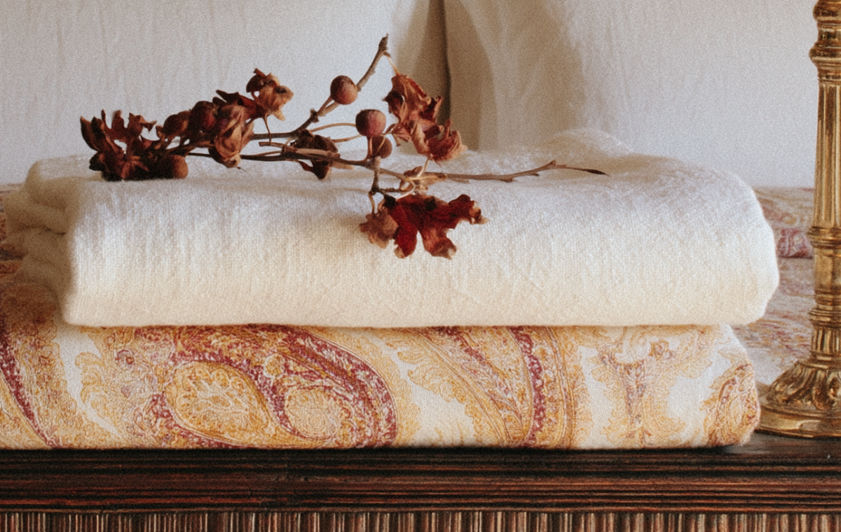 Linen blanket layering with neutral throws, vintage paisley quilts, and dried botanical stems on a wooden surface.