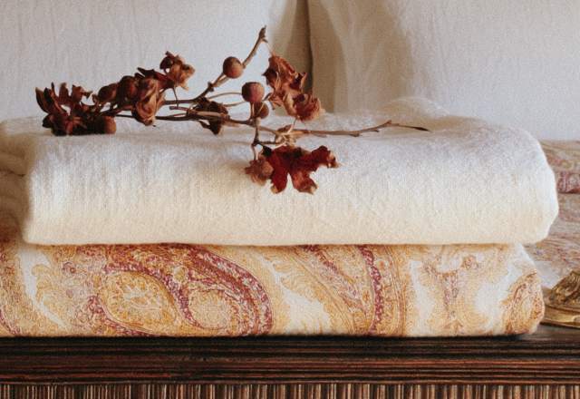 Linen blanket layering with neutral throws, vintage paisley quilts, and dried botanical stems on a wooden surface.
