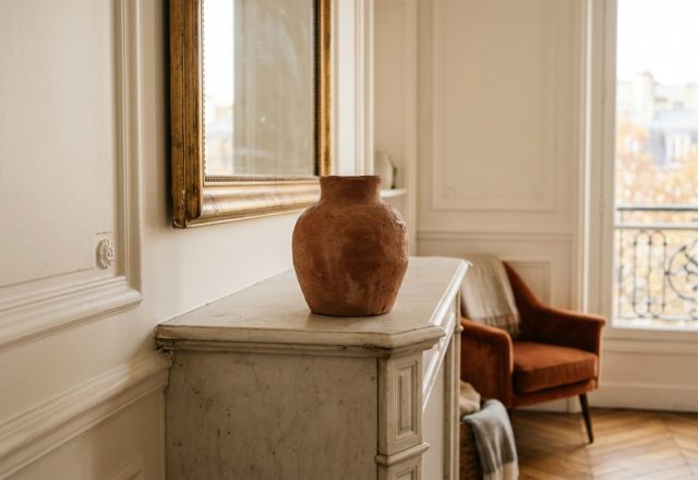 Minimalist mantel styling featuring a terracotta vase on a white marble fireplace with gold mirror and French windows in background