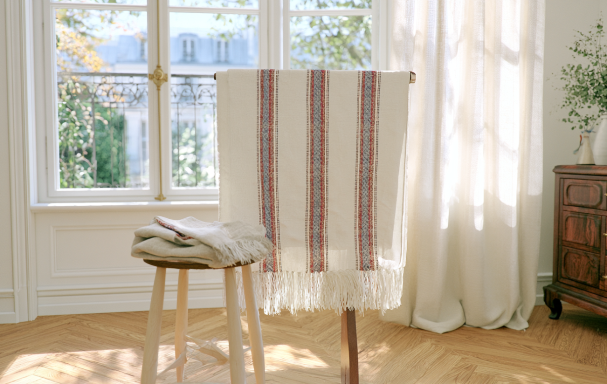 Natural textile styling featuring a striped linen throw and folded fabrics on a wooden stool near a sunlit window.