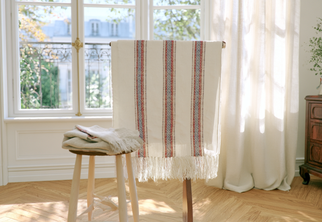 Natural textile styling featuring a striped linen throw and folded fabrics on a wooden stool near a sunlit window.