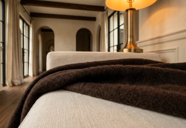 Close-up of a dark brown throw blanket on a neutral linen bench inside a styled living room with archways.