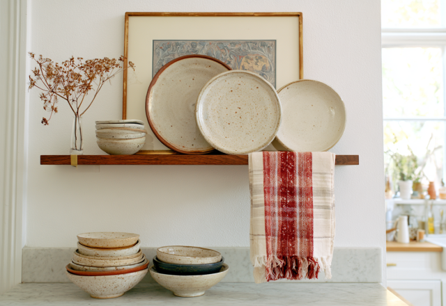 Open kitchen shelving styled with handmade ceramic plates, bowls, framed art, and a hanging red patterned towel.