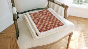 A crimson patterned quilt folded neatly on a cream vintage armchair in a bright living space.