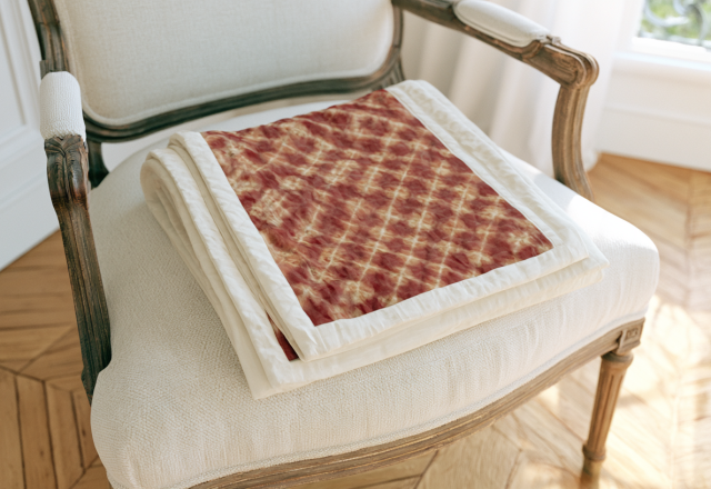 A crimson patterned quilt folded neatly on a cream vintage armchair in a bright living space.