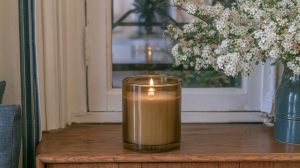 Amber glass scented candle burning on a wooden window sill next to white blossoms