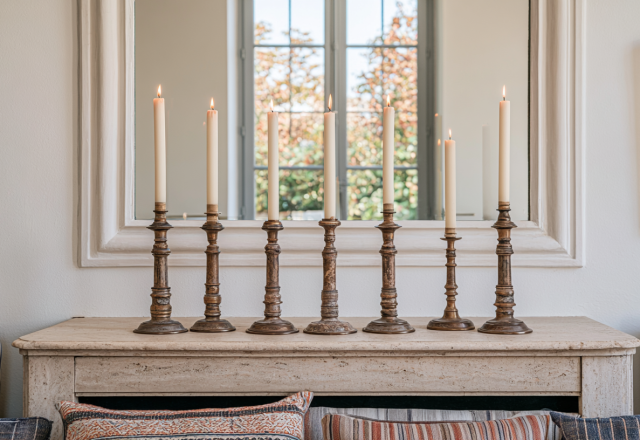 A sleek mantel candle display featuring seven antique brass candlesticks with lit white taper candles on a stone fireplace ledge.