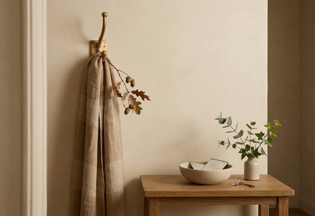 Minimalist entryway decor featuring a brass wall hook with draped throw, oak table, and ceramic catchall bowl.