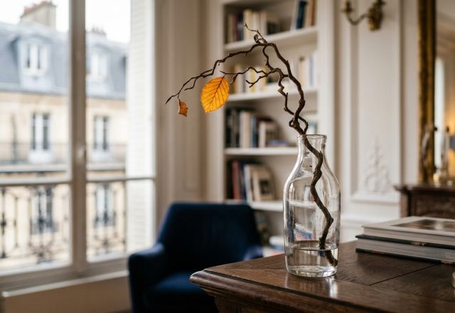 Minimalist home styling featuring a curved natural branch in a glass vase on a wooden desk near a tall window.