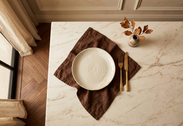 Minimalist tablescape featuring a white ceramic plate, dark linen napkin, and brass cutlery on a marble countertop.
