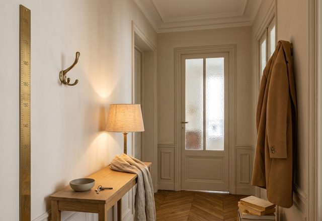 Narrow hallway decor showing slim console table, warm small lamp, brass growth ruler, and chevron parquet floor.