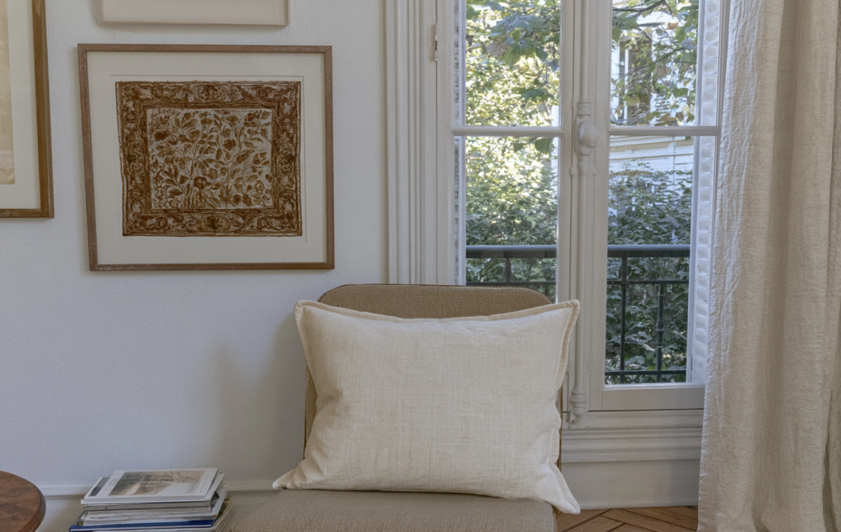 Neutral window seat decor featuring a linen pillow, framed botanical art, and a stack of books near a sunny French window.