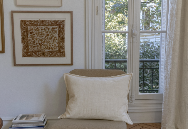Neutral window seat decor featuring a linen pillow, framed botanical art, and a stack of books near a sunny French window.