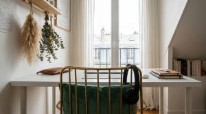 A clean small home office setup featuring a white desk, green velvet chair, and natural light from a window.