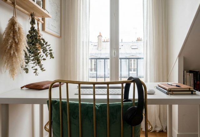 A clean small home office setup featuring a white desk, green velvet chair, and natural light from a window.