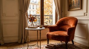 Cozy Parisian living room decor featuring a rust velvet armchair, marble side table, and classic wainscoting against a window view.