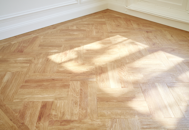 Close up view of natural oak parquetry wood flooring illuminated by afternoon sunlight next to white baseboards.