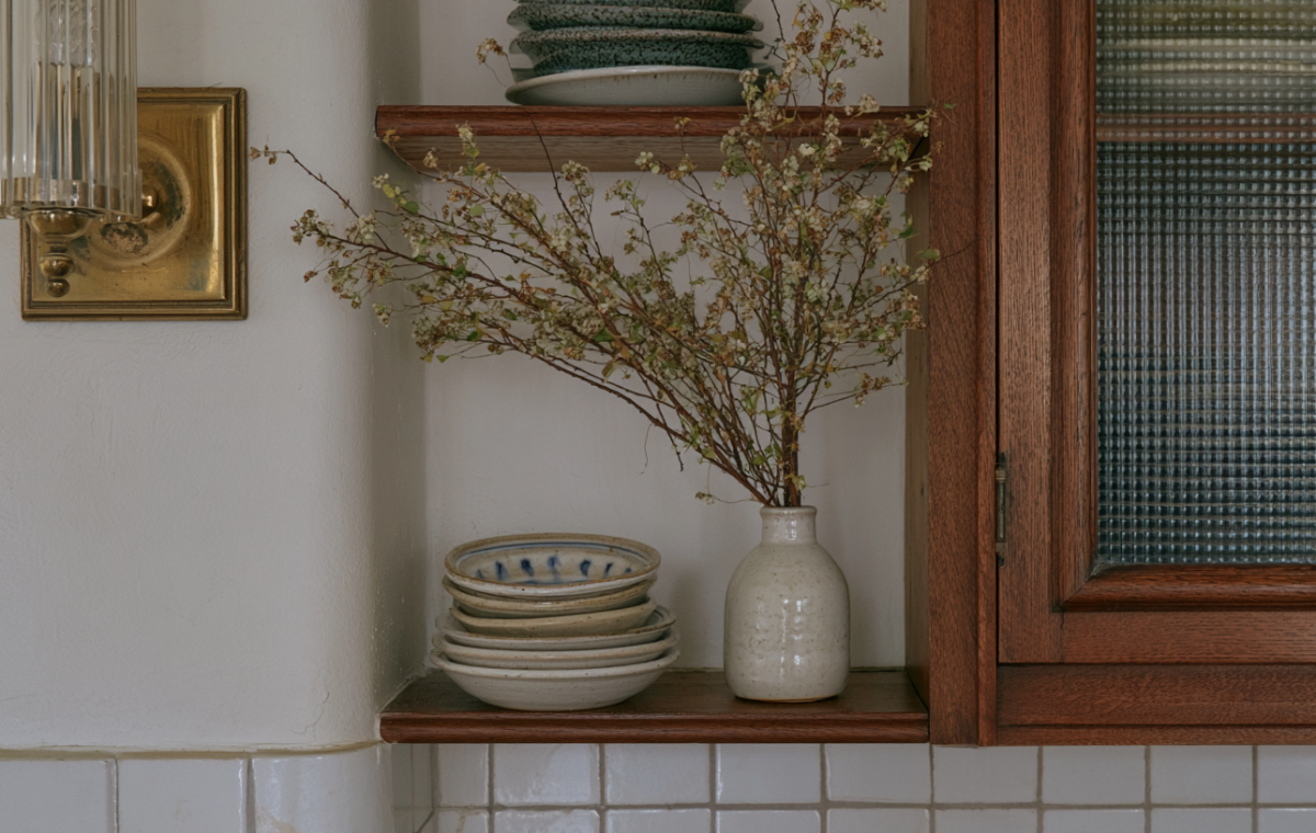 Wooden kitchen open shelving styled with ceramic plates, a vase with dried branches, and an adjacent glass pantry cabinet.