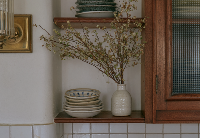 Wooden kitchen open shelving styled with ceramic plates, a vase with dried branches, and an adjacent glass pantry cabinet.