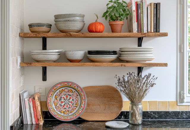Open kitchen shelving styled with ceramic bowls, plates, books, and a small potted herb plant.