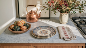 Kitchen countertop styling featuring a copper kettle, wooden bowl of gourds, and stacked ceramic plates on marble.