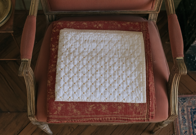 An antique chair cushion with a white quilted cover resting on a red damask vintage seat in a historic room setting.