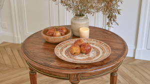 A styled round wooden coffee table featuring a ceramic plate of pears, a wooden bowl, a vase with dried botanicals, and a lit candle.