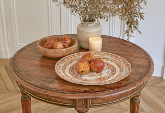 A styled round wooden coffee table featuring a ceramic plate of pears, a wooden bowl, a vase with dried botanicals, and a lit candle.