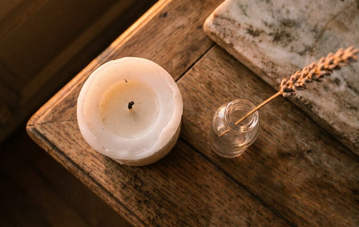 White pillar candle and dried botanical stem in a glass bottle on a rustic wooden table.