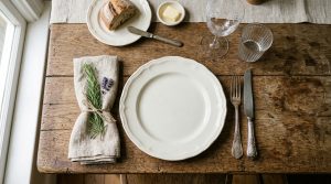 A rustic table setting featuring a white ceramic plate, linen napkin with rosemary, and vintage silverware on a wooden table.