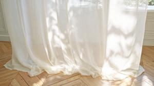 Sunlit sheer linen curtains pooling onto herringbone wood flooring in a bright living room.