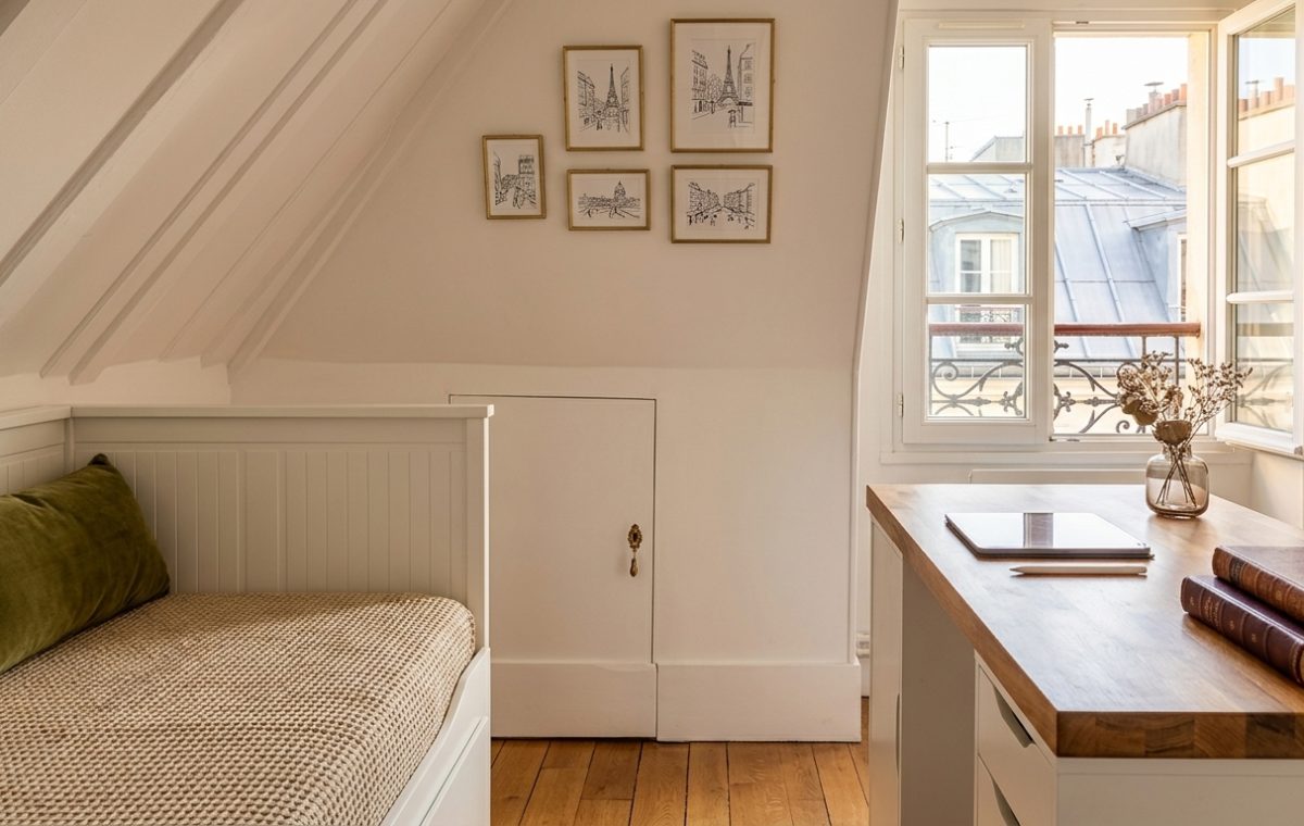 Cozy attic bedroom office featuring a white daybed, wooden desk, and sloped ceilings under natural light.