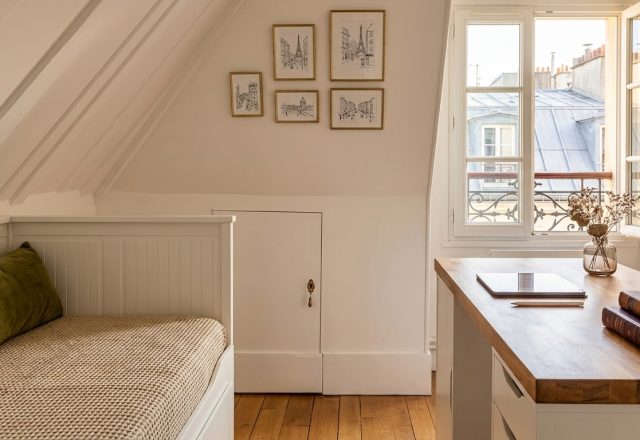 Cozy attic bedroom office featuring a white daybed, wooden desk, and sloped ceilings under natural light.