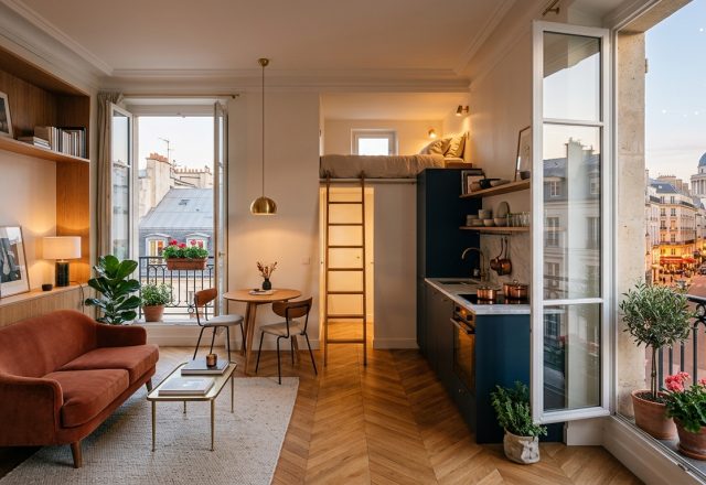 Bright small studio apartment design featuring a loft bed, navy kitchen, and rust-colored sofa overlooking a city street.