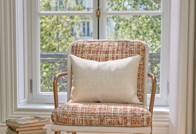 Cozy reading nook featuring a textured plaid armchair with a neutral linen cushion and stacked books near a sunny window.